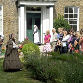 Tour group stood watching two costumed characters perform in garden of brick built house with green door